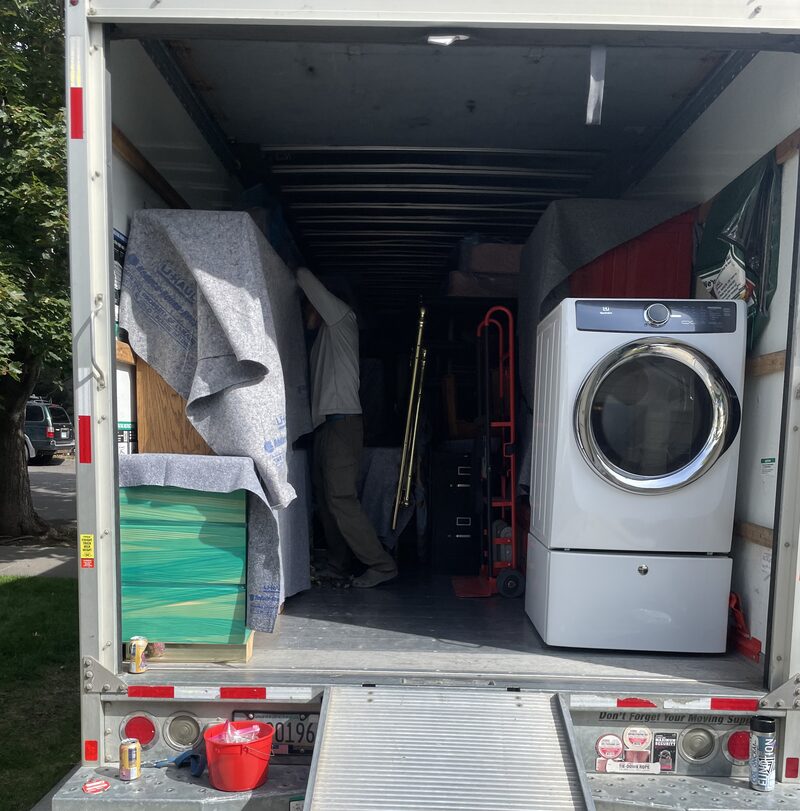 Loaded moving truck with the ramp down, holding a washer, hand truck and pad-wrapped furniture
